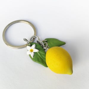 Puede incluir: Un llavero artesanal con un lim&oacute;n amarillo brillante, hojas verdes y una flor blanca con centro amarillo. El lim&oacute;n est&aacute; unido a un llavero plateado, perfecto para llaves o bolsos.