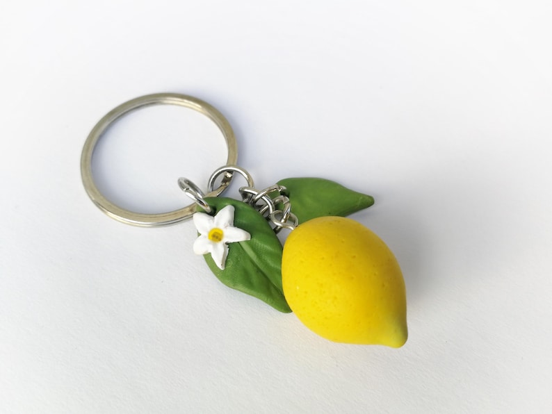Puede incluir: Un llavero artesanal con un lim&oacute;n amarillo brillante, hojas verdes y una flor blanca con centro amarillo. El lim&oacute;n est&aacute; unido a un llavero plateado, perfecto para llaves o bolsos.