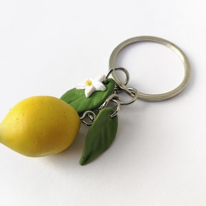 Puede incluir: Un llavero artesanal con un lim&oacute;n amarillo, hojas verdes y una flor blanca. El lim&oacute;n tiene una superficie texturizada. El dije est&aacute; unido a un llavero plateado, perfecto para llaves o bolsos.