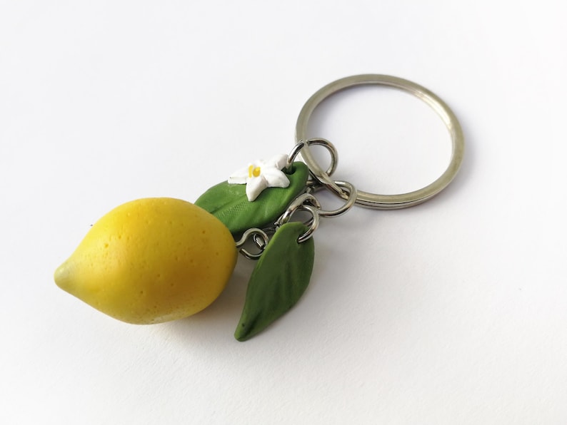 Puede incluir: Un llavero artesanal con un lim&oacute;n amarillo, hojas verdes y una flor blanca. El lim&oacute;n tiene una superficie texturizada. El dije est&aacute; unido a un llavero plateado, perfecto para llaves o bolsos.