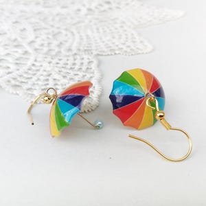 Puede incluir: Un par de pendientes de paraguas coloridos. Cada pendiente presenta un diseño de paraguas en miniatura con un arcoíris de colores, incluyendo rojo, naranja, amarillo, verde, azul y morado. Los pendientes tienen ganchos dorados y una pequeña cuenta azul.