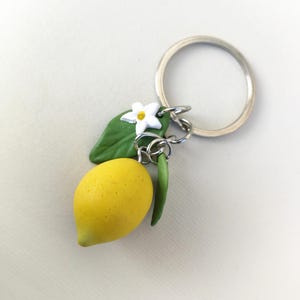 Puede incluir: Un llavero artesanal con un lim&oacute;n amarillo brillante con una hoja verde, una flor blanca y un tallo verde. El llavero est&aacute; unido a un llavero de metal plateado.