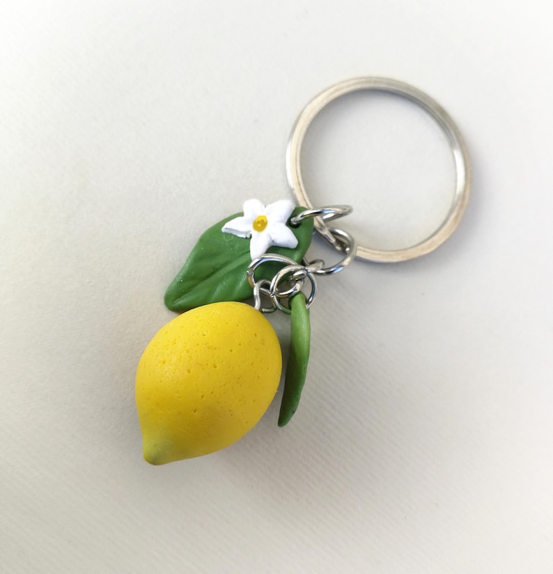 Puede incluir: Un llavero artesanal con un lim&oacute;n amarillo brillante con una hoja verde, una flor blanca y un tallo verde. El llavero est&aacute; unido a un llavero de metal plateado.