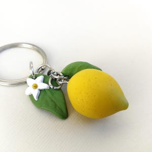 Puede incluir: Un llavero hecho a mano con un dije de lim&oacute;n amarillo, hojas verdes y una flor blanca. El lim&oacute;n mide aproximadamente 4 cm de largo. El llavero incluye un anillo y una cadena plateados.