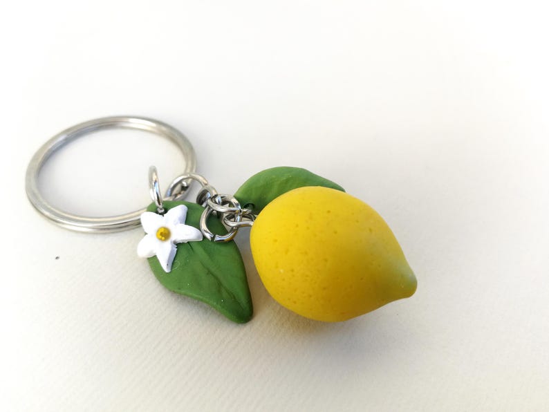 Puede incluir: Un llavero hecho a mano con un dije de lim&oacute;n amarillo, hojas verdes y una flor blanca. El lim&oacute;n mide aproximadamente 4 cm de largo. El llavero incluye un anillo y una cadena plateados.