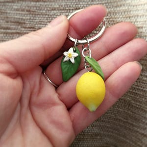 Puede incluir: Un llavero hecho a mano con un lim&oacute;n en miniatura, una flor blanca y hojas verdes. El lim&oacute;n es de color amarillo brillante y las hojas son de un verde vibrante. El llavero est&aacute; unido a un anillo plateado.