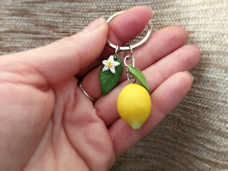 Puede incluir: Un llavero hecho a mano con un lim&oacute;n en miniatura, una flor blanca y hojas verdes. El lim&oacute;n es de color amarillo brillante y las hojas son de un verde vibrante. El llavero est&aacute; unido a un anillo plateado.