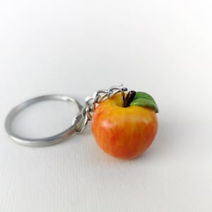 Könnte beinhalten: Ein roter und gelber Apfel-Schlüsselanhänger mit einem grünen Blatt. Der Apfel ist an einem silbernen Schlüsselring befestigt.