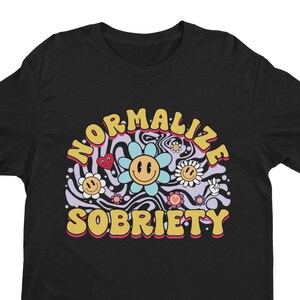 Puede incluir: Camiseta negra con un diseño psicodélico que incluye el texto "Normalizar la sobriedad" en letras amarillas con contorno rosa. El diseño incluye una cara sonriente con un corazón y flores.