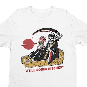May include: White t-shirt with a graphic of a skeleton holding a rose and a scythe, sitting in a coffin. The text "STILL SOBER BITCHES" is printed below the image.