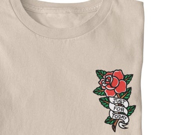 May include: A beige t-shirt with a red rose and green leaves embroidered on the front. The rose has a banner that reads "Just For Today".