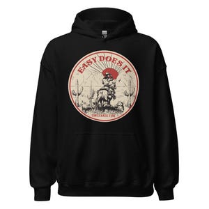 May include: Black hoodie featuring a vintage-style graphic of a cowboy riding a horse in front of a sunset. The graphic is in a circle with the text "Easy Does It" and "Time Takes Time" below it.