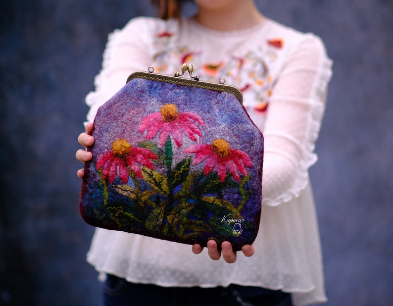 May include: A purple felt clutch bag with a floral design featuring pink and yellow flowers. The bag has a metal clasp and a small metal tag with the word "Kraynvy" on it.