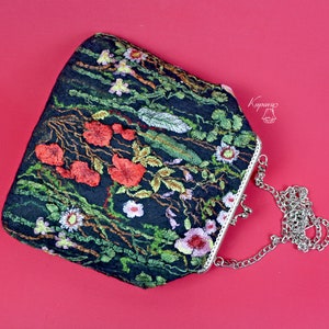 May include: A black velvet clutch bag with a floral embroidery design featuring red, green, and white flowers. The bag has a silver chain strap and a silver clasp.