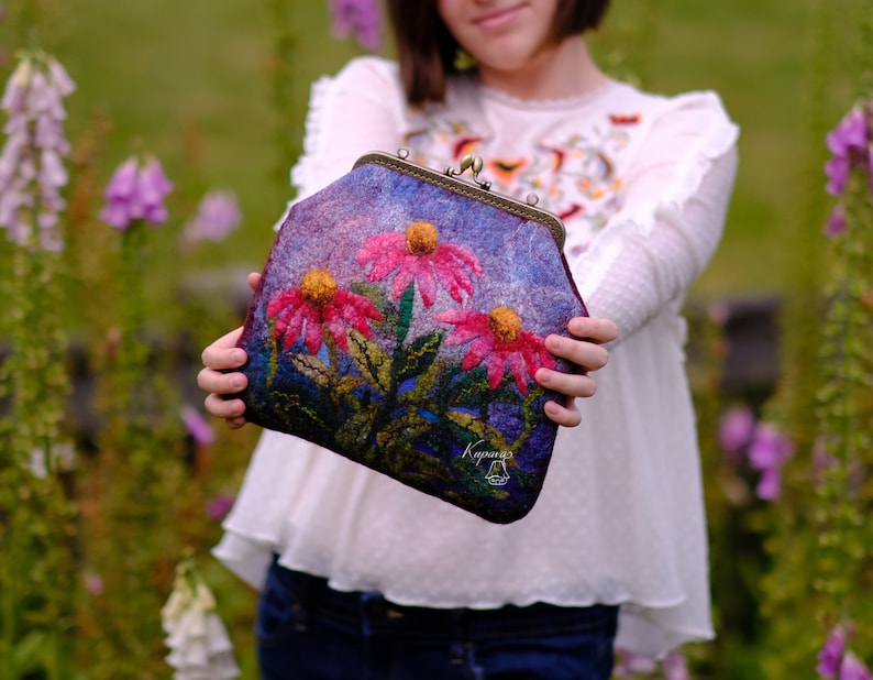 May include: A purple felt clutch bag with a floral design featuring pink and yellow flowers. The bag has a metal clasp and the text "Kayana" is embroidered on the front.
