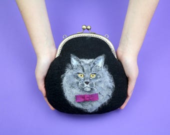 May include: A black coin purse with a metal kiss-lock clasp featuring a needle-felted gray cat portrait on the front. The cat has yellow eyes and wears a small, bright magenta bow tie. The purse has a rounded shape and a textured fabric surface.
