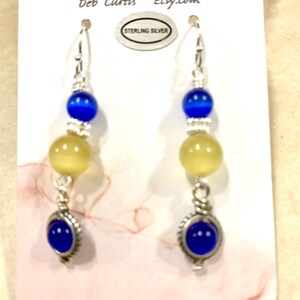 May include: A pair of dangle earrings with a silver hook, featuring blue and yellow beads. The earrings have a blue, oval-shaped stone at the bottom. The card behind the earrings reads "BEADazzlingbydeb" and "Etsy.com".