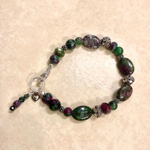 May include: A bracelet featuring polished oval and round ruby zoisite beads in shades of deep green and magenta. The beads are interspersed with textured silver-toned metal spacers. It includes a silver-toned toggle clasp and a small heart charm.