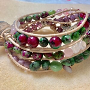 May include: Wrap bracelet featuring a mix of ruby zoisite, rose quartz, and amethyst beads. The beads are strung on a light brown leather cord, with a silver-toned button clasp. The bracelet is designed to wrap multiple times around the wrist.