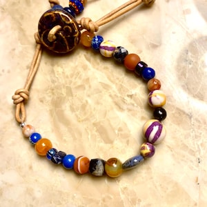 May include: A beaded bracelet featuring a variety of multicolored spherical beads in shades of blue, orange, purple, and brown, strung on a tan leather cord. The bracelet includes a large, dark brown ceramic button closure with a spiral design.
