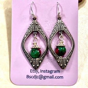 May include: A pair of silver-toned earrings with a diamond-shaped frame featuring intricate scrollwork. Each earring has a green and red gemstone bead suspended within the frame. The earrings are displayed on a lavender card with Etsy, Instagram, and an email address.