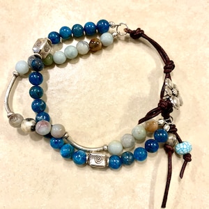 May include: A multi-strand bracelet with blue, green, and brown beads, silver accents, and a brown leather cord with a silver flower charm.