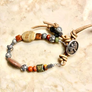 May include: A wrap bracelet featuring tan leather cord, a circular silver-toned metal button with a dragonfly motif, and an assortment of beads including smooth stone, textured metal, and square-shaped jasper in shades of brown, gray, and green.