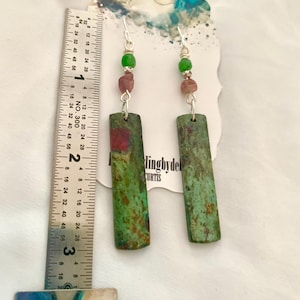 May include: A pair of green and pink earrings with a rectangular green stone and silver hooks. The earrings are approximately 2 inches long.