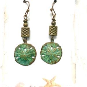 May include: A pair of dangle earrings with a beach theme. The earrings feature a green sand dollar pendant with a bronze-colored metal accent. The earrings have a decorative square bead and a curved ear wire. The text "BEADAZZLINGbydeb" is at the top.