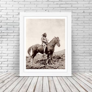 Op de afbeelding: Een zwart-witfoto van een indiaanse man die op een paard rijdt. De man draagt een verenhoofdtooi en houdt een speer vast. Het paard is bruin en heeft een witte ster op zijn voorhoofd.