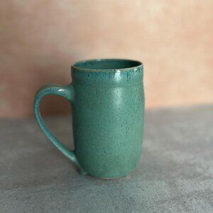 Puede incluir: Una taza de cerámica hecha a mano en color verde mar. La taza tiene un cuerpo redondeado, un asa curvada y una superficie ligeramente texturizada. El borde de la taza tiene un esmalte de color verde azulado más oscuro. La taza está sobre un fondo neutro.