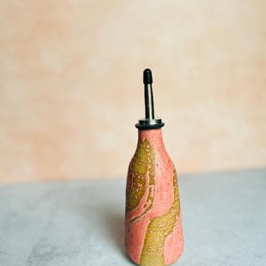 Könnte beinhalten: Eine rosa und braune Keramikflasche für Öl und Essig mit einem schwarzen Ausgießer.
