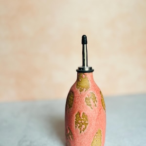 Può includere: Una bottiglia di ceramica rosa per olio e aceto con un design a foglia d'oro. La bottiglia ha un beccuccio nero.