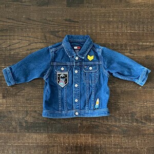 May include: A blue denim jacket with a yellow chevron patch, a space explorer patch, and a rocket patch. The jacket has a button closure and two pockets.