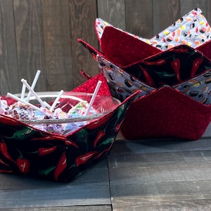 May include: Two fabric bowls, one red with white polka dots and one black with red chili peppers, are filled with candy. The red bowl is stacked on top of another fabric bowl with a sushi pattern.