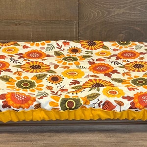May include: A white oval-shaped baking dish cover with a yellow border. The cover is decorated with a floral pattern featuring sunflowers, leaves, and other fall-themed elements in shades of orange, yellow, brown, and green.