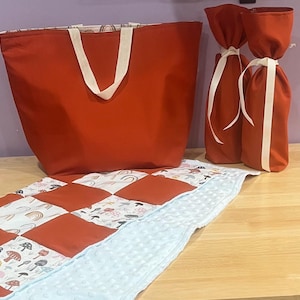 May include: A large, red tote bag with cream-colored handles sits next to two red wine bottle bags tied with cream ribbons. A patchwork quilt with red squares and a light blue, textured border is in the foreground.