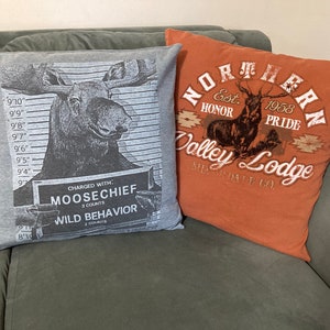 May include: Two decorative throw pillows. One pillow is gray with a black and white image of a moose with the text "CHARGED WITH: MOOSECHIEF. 3 COUNTS WILD BEHAVIOR 2 COUNTS". The other pillow is orange with the text "NORTHERN Est 1958 HONOR PRIDE Valley Lodge SUR DATE CO."