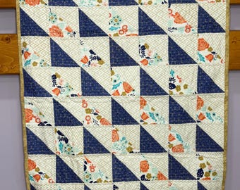 May include: A rectangular quilt featuring a half-square triangle pattern. The design alternates between dark blue fabric with a gold geometric print and cream fabric with a floral pattern in orange, teal, and gold. It has a gold-toned border.