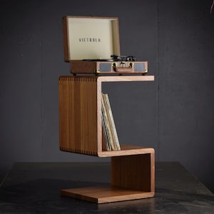 May include: A brown Victrola record player sits atop a wooden side table with a unique, zig-zag design. The table has a lower shelf holding a stack of vinyl records. The record player's case is open, revealing the turntable.