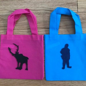 Puede incluir: Dos peque&ntilde;as bolsas de mano con asas de tela. La bolsa de la izquierda es rosa con una silueta negra de un reno. La bolsa de la derecha es azul con una silueta negra de una persona.