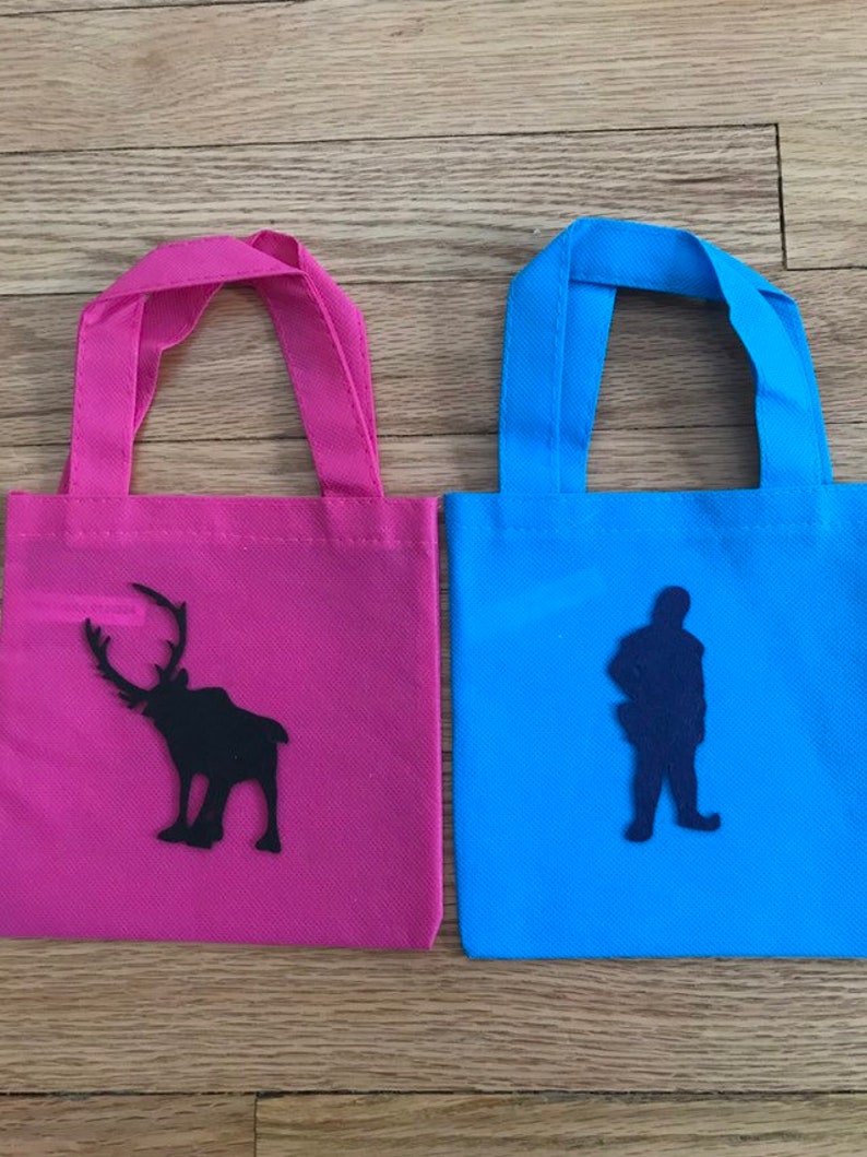 Puede incluir: Dos peque&ntilde;as bolsas de mano con asas de tela. La bolsa de la izquierda es rosa con una silueta negra de un reno. La bolsa de la derecha es azul con una silueta negra de una persona.