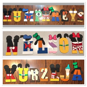 May include: Three wooden names, HUMPHREYS, NELSON, and KURZEJA, are spelled out in large, colorful letters. Each letter is decorated with a different Disney character, such as Mickey Mouse, Donald Duck, and Goofy.