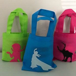 Puede incluir: Tres peque&ntilde;as bolsas de tela reutilizables con diferentes dise&ntilde;os. La primera bolsa es verde con una silueta rosa de una mujer. La segunda bolsa es azul con una silueta blanca de una mujer. La tercera bolsa es rosa con una silueta negra de un reno.