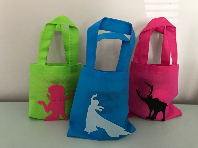 Puede incluir: Tres peque&ntilde;as bolsas de tela reutilizables con diferentes dise&ntilde;os. La primera bolsa es verde con una silueta rosa de una mujer. La segunda bolsa es azul con una silueta blanca de una mujer. La tercera bolsa es rosa con una silueta negra de un reno.