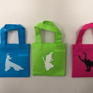 Puede incluir: Tres bolsas de mano peque&ntilde;as con diferentes dise&ntilde;os. La bolsa azul tiene una silueta blanca de una mujer, la bolsa verde tiene una silueta blanca de un hada y la bolsa rosa tiene una silueta negra de un reno.