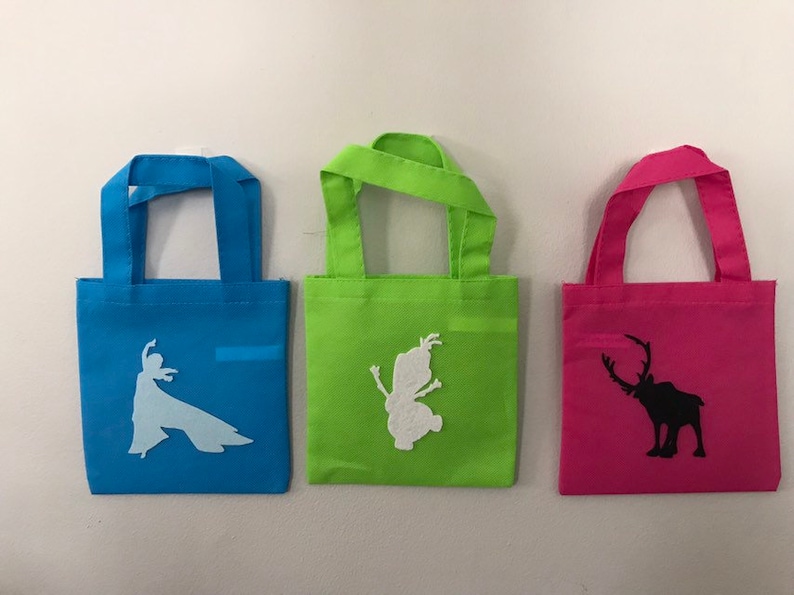 Puede incluir: Tres bolsas de mano peque&ntilde;as con diferentes dise&ntilde;os. La bolsa azul tiene una silueta blanca de una mujer, la bolsa verde tiene una silueta blanca de un hada y la bolsa rosa tiene una silueta negra de un reno.