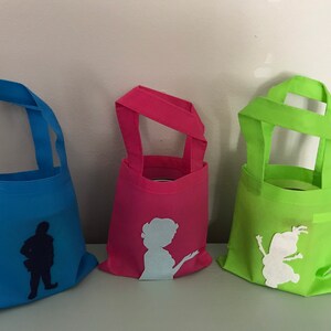 Puede incluir: Tres peque&ntilde;as bolsas de tela reutilizables con diferentes siluetas de personajes. La bolsa de la izquierda es azul con una silueta azul oscuro de un hombre. La bolsa del medio es rosa con una silueta azul claro de una mujer. La bolsa de la derecha es verde con una silueta blanca de un personaje de dibujos animados.