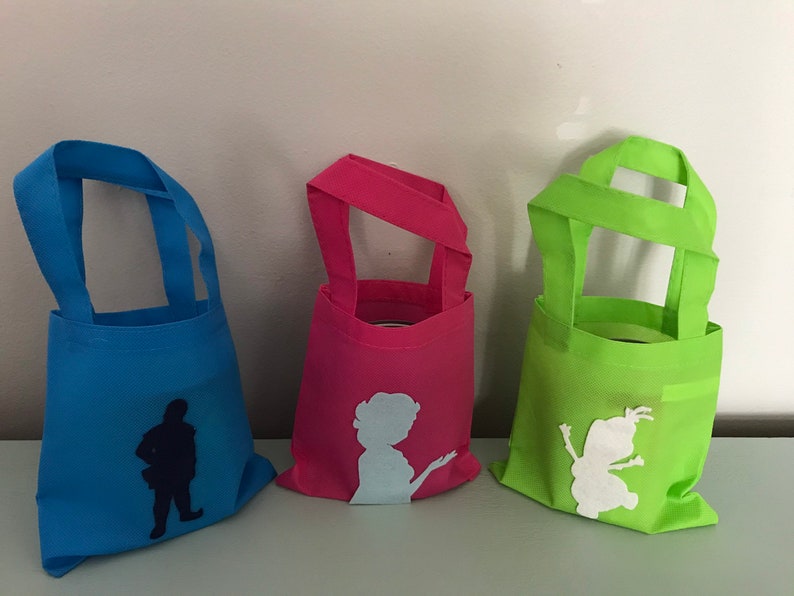 Puede incluir: Tres peque&ntilde;as bolsas de tela reutilizables con diferentes siluetas de personajes. La bolsa de la izquierda es azul con una silueta azul oscuro de un hombre. La bolsa del medio es rosa con una silueta azul claro de una mujer. La bolsa de la derecha es verde con una silueta blanca de un personaje de dibujos animados.