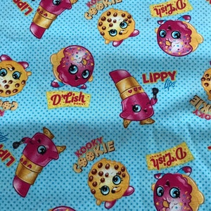 May include: A blue fabric with a repeating pattern of cartoon characters. The characters are a cookie, a donut, and a lipstick. The characters have faces and are wearing different outfits. The fabric has a white polka dot pattern.